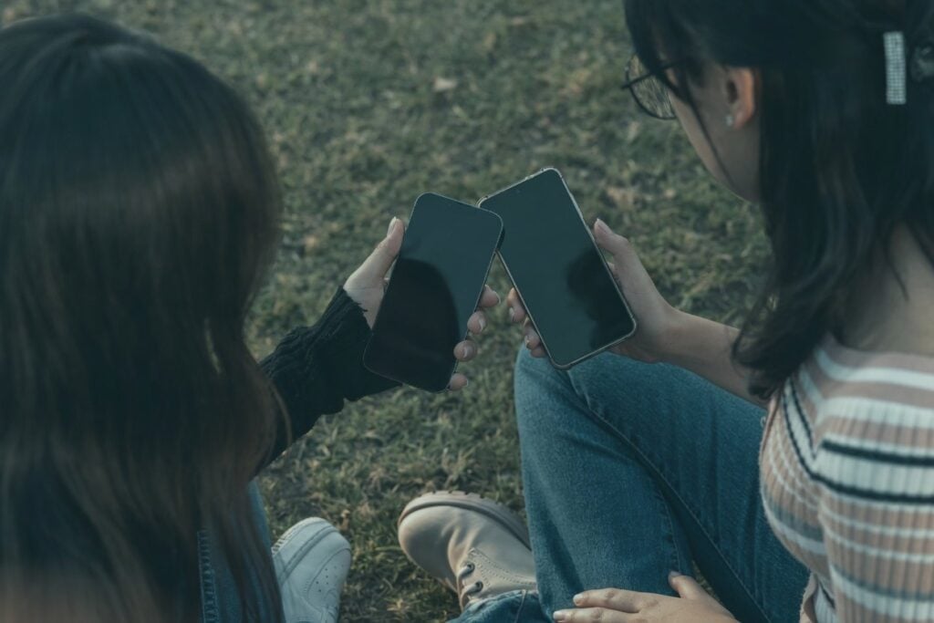 Two women sit outdoors holding smartphones, suggesting love bombing dating apps.