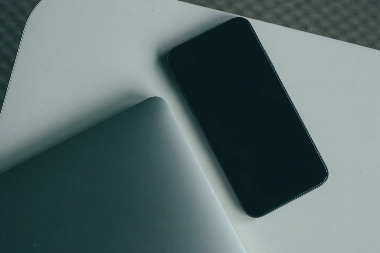 A black smartphone sits next to a silver laptop on a white surface.