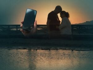 Silhouetted couple near water at sunset with a hand holding a smartphone, people search to find ex.