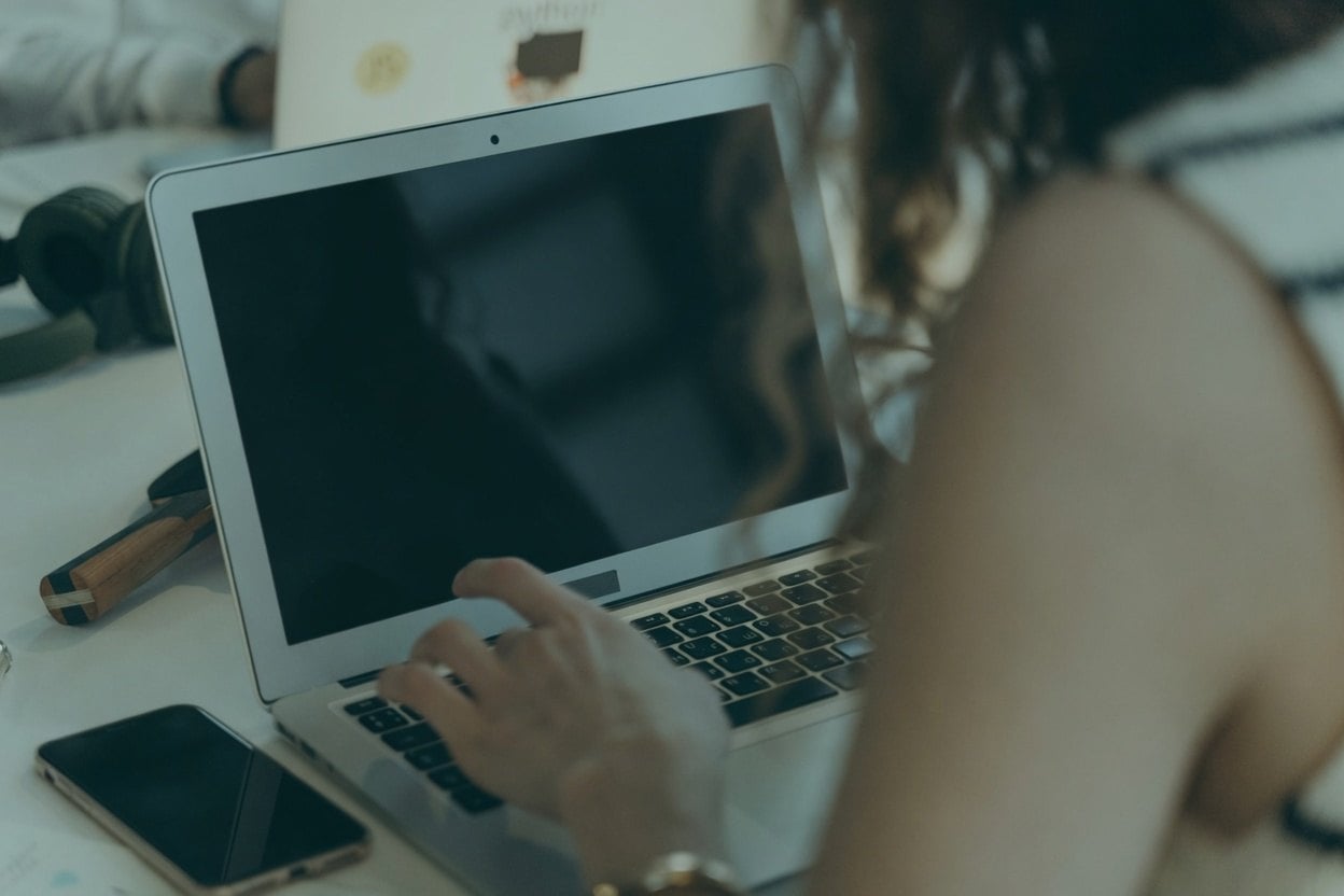 How To Use LinkedIn To Find An Address 1 A woman is typing on a laptop with a cell phone nearby.