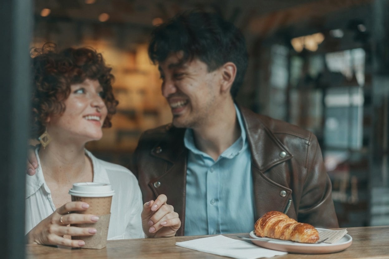 Funny First Date Questions 1 A man and woman laughing together at a cafe, with a croissant on the table