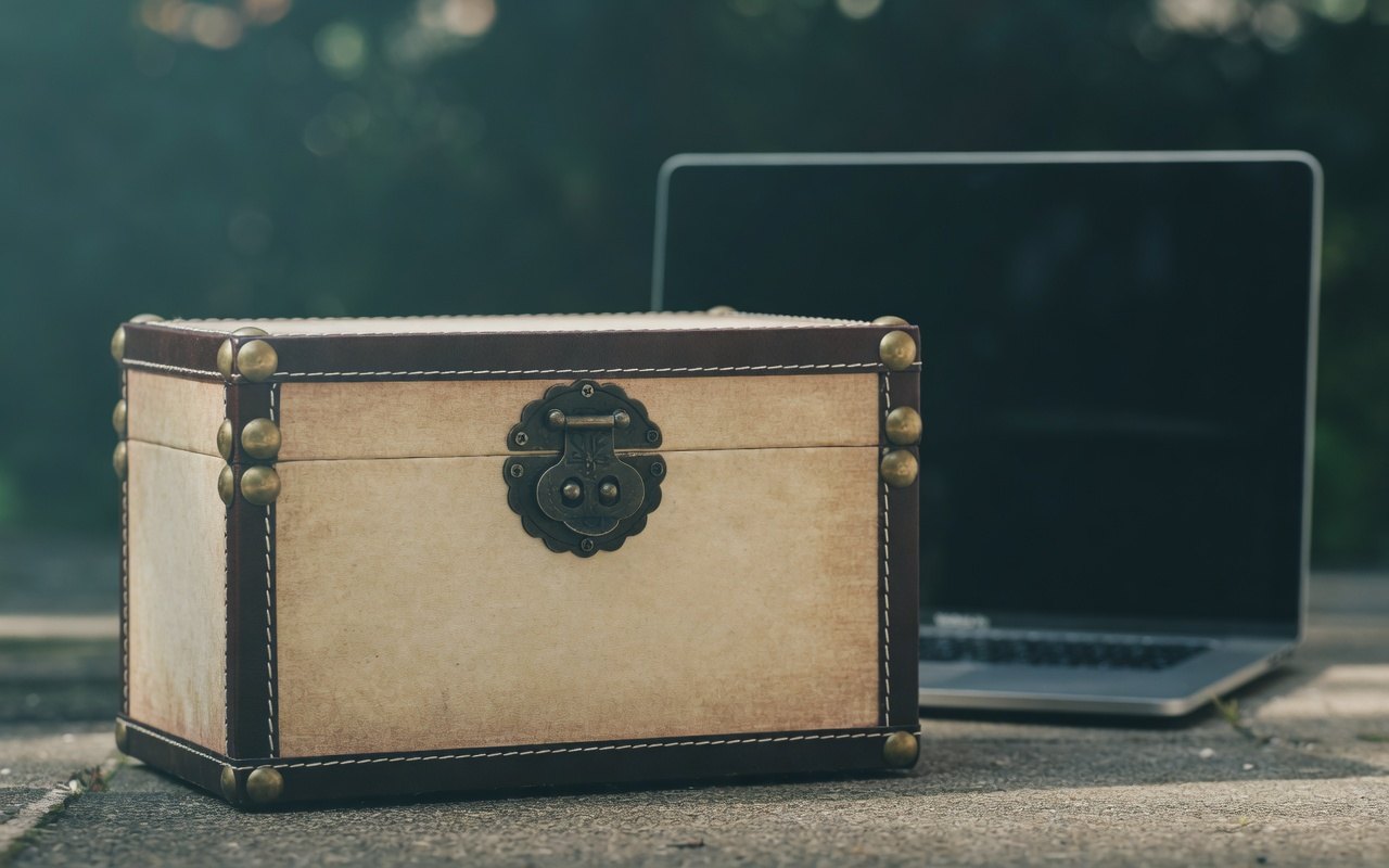 A vintage trunk with a metal lock sits next to an open laptop on a concrete surface.