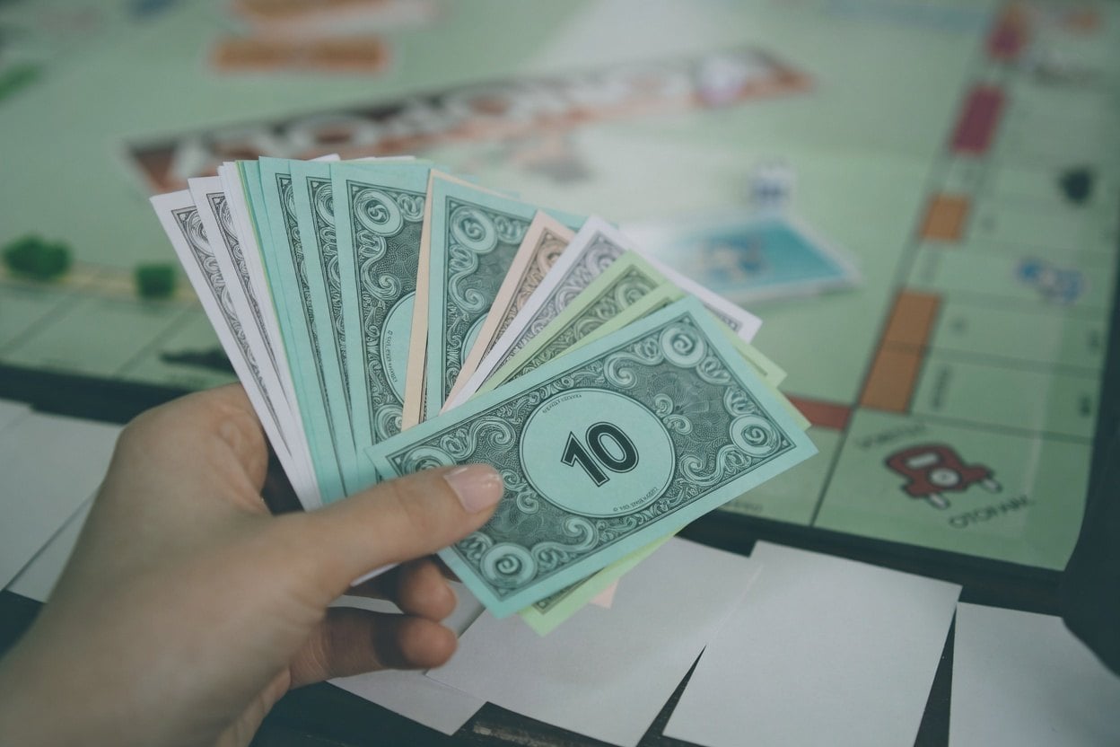 A hand holding a fan of $10 bills with a Monopoly board in the background.