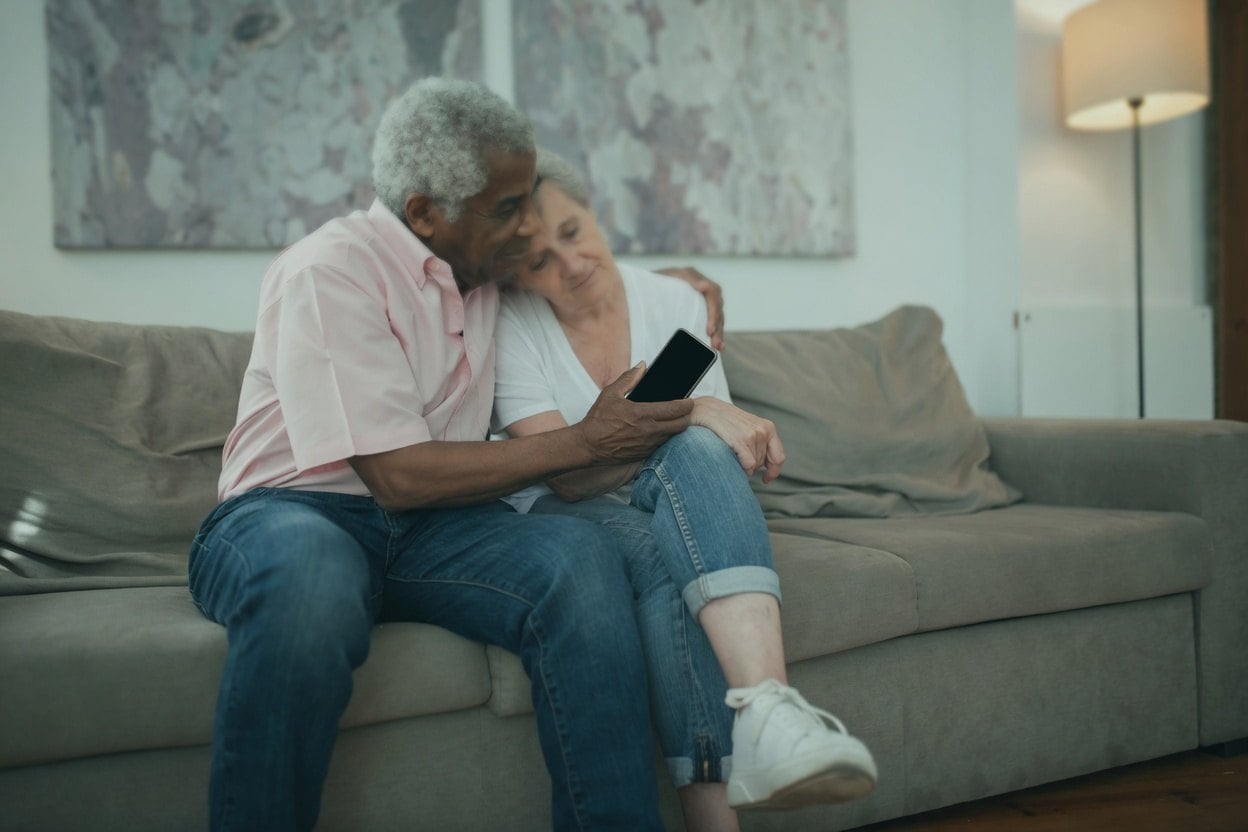 A man and woman sitting on a couch, one holding a phone, with a lamp in the background