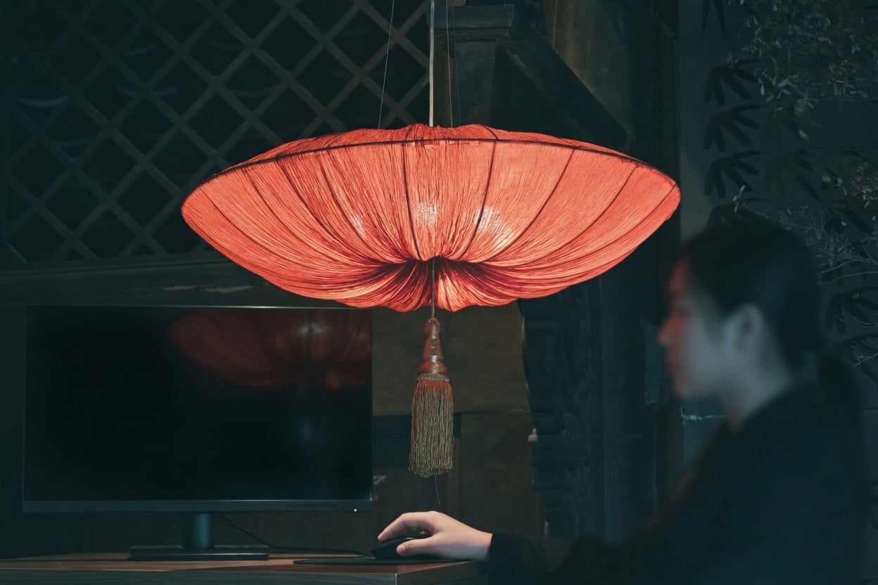 A woman sits at a desk with a red lantern hanging above her, illuminating her workspace.